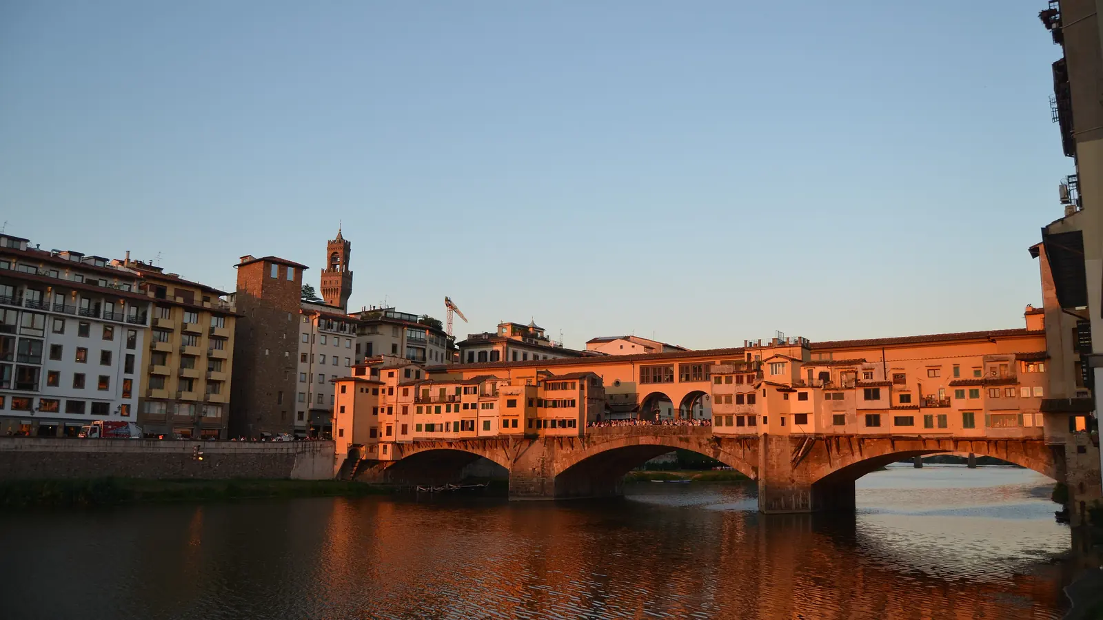 The Ponte Vecchio at sunset, its shops glowing orange above the Arno, with the tower of the Palazzo Vecchio behind