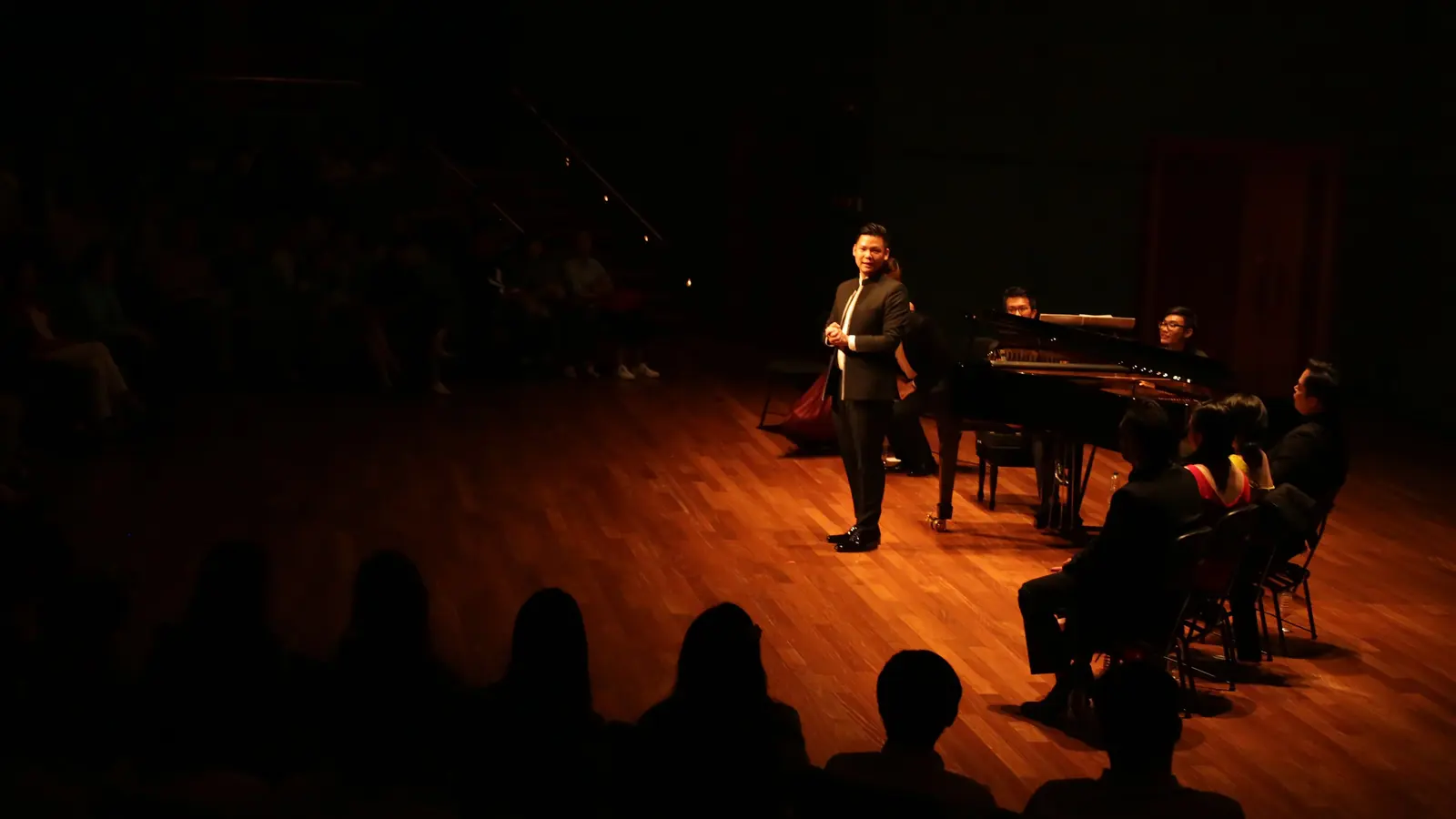 A tenor stands in a pool of warm stage light beside a grand piano while a darkened audience listens; a tenor recital in another hall, not the Florence concert
